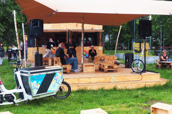 Foto vom mobilen Kultur·laden KommVorZone im Anna·park. Auf einer Wiese steht ein Zelt. Unter dem Zelt steht eine Bühne aus Holz. vor der Bühne stehen Holz·würfel zum Sitzen.