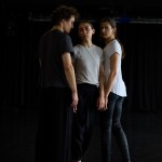 drei Menschen stehen dicht beeinander und schauen in die Kamera, es sind zwei Tänzerinnen und ein Tänzer, sie berühren sich leicht an den Händen, der Tänzer schaut nicht in die Kamera. Sie sind barfuß.