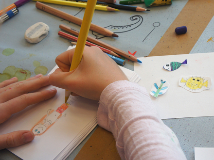 Foto von 2 Händen von einem Kind. Die rechte Hand hält einen gelben Bunt·stift. Das Kind zeichnet mit Bunt·stiften., Bild © KinderKunstRaum / Stadt Nürnberg