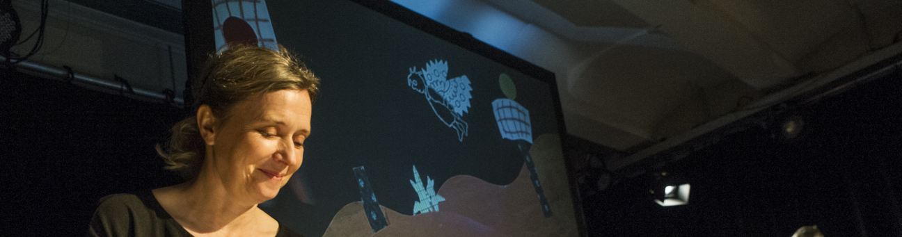 Frau an einem Legebrett setzt Figuren um, die als Projekton an einer leinwand zu sehen sind zum Beispiel Turm, Engel und Pflanzen. Im Hintergrund ein Mann am Akkordeon mit Notenständer.