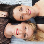 Eine blonde und eine dunkelhaarige Frau liegen auf einer weißen Fläche und lächeln in die Kamera