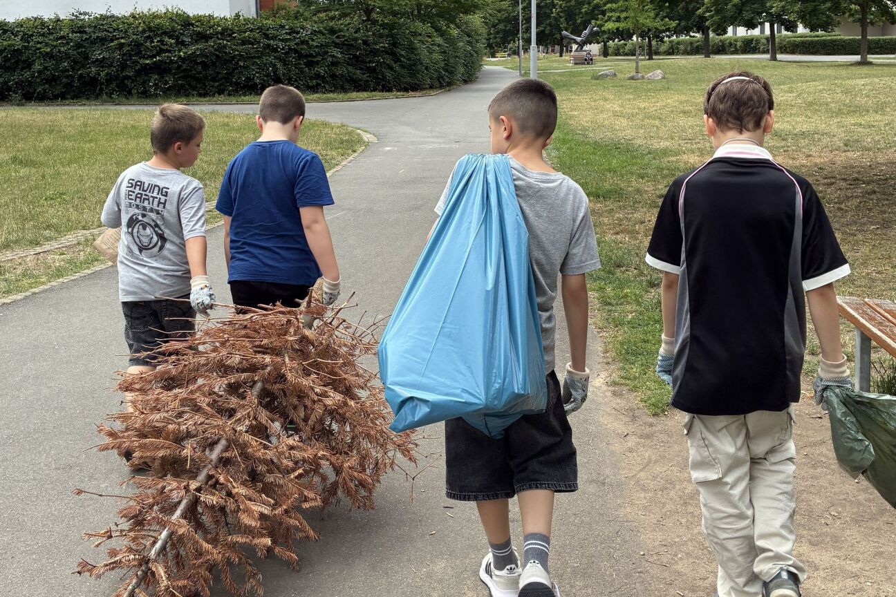 Vier Kinder laufen ausgestattet mit Müllsäcken umher, einer hat einen vertrockneten Tannenbaum im Schlepptau, während ein anderer einen blauen Müllsack trägt. Alle tragen Müllhandschuhe zum Müllsammeln.