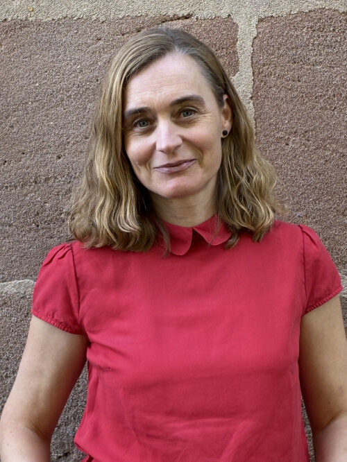 © Kulturladen Röthenbach / Stadt Nürnberg Eine Frau mit blonden, schulterlangen und leicht lockigen Haaren steht vor einer Sandsteinwand und lächelt. Sie trägt eine rote Bluse und schwarze dezente Ohrringe. © Kulturladen Röthenbach / Stadt Nürnberg