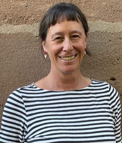 Eine Frau mit braunen Haaren und einem engen, gestreiften Oberteil steht vor einer Sandsteinwand und lächelt. Sie trägt dezente rot-rosa Ohrringe.
