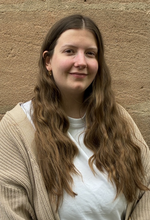 Eine Junge Frau mit dunkelblonden Haaren, einem weißen T-Shirt und einer beigen Strickjacke