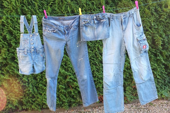 Eine Kinderlatzhose, ein Kinderrock und zwei lange Hosen, alle aus Jeansstoff, hängen an einer Wäscheleine vor einem grünen Busch