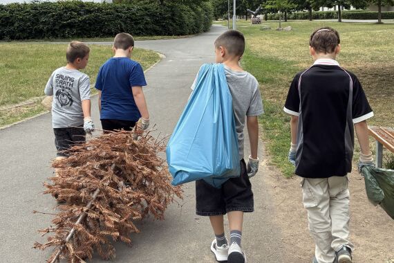 Vier Kinder laufen ausgestattet mit Müllsäcken umher, einer hat einen vertrockneten Tannenbaum im Schlepptau, während ein anderer einen blauen Müllsack trägt. Alle tragen Müllhandschuhe zum Müllsammeln.