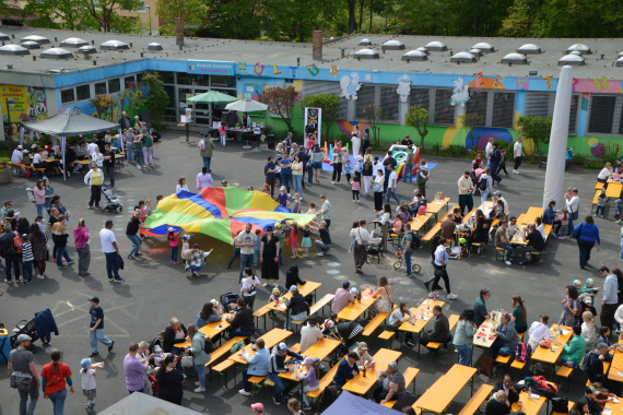Auf einem großen Platz sind viele Familien anwesend. Sie sitzen an Biertischen oder laufen herum. In der Mitte wird von Kindern ein großes Buntes Tuch gehalten.