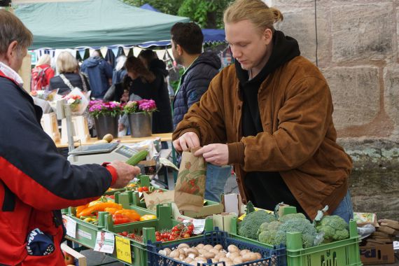 Ein Mann mit brauner Jacke und blonden Haaren steht mit einer Papiertüte hintereinem Verkaufsstand mit Gemüse. Ein anderer Mann steht ihm gegenüber und reicht ihm eine Salatgurke.