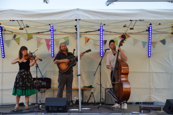 Frau mit Geige, Mann mit Gitarre und Mann mit Kontrabass spielen zusammen auf einer Bühne in einem Pavillon