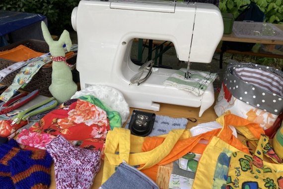 Eine weiße Nähmaschine sthet auf einem Tisch. Um sie herum liegen verschiedene kleine Taschen, Mützen und Schlüsselbänder aus buntem Stoff und kleine Stricksocken.