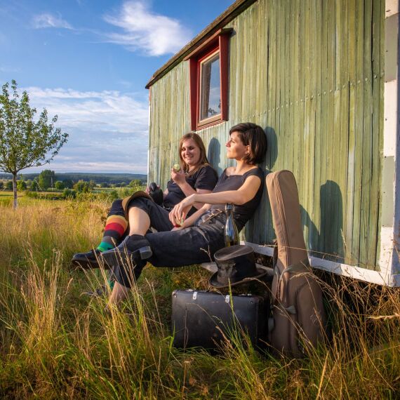 Zwei Frauen sitzen vor einem Bauwagen und genießen die Sonne. Ein Gitarrenkoffer und weitere Taschen liegen vor ihnen im Gras.