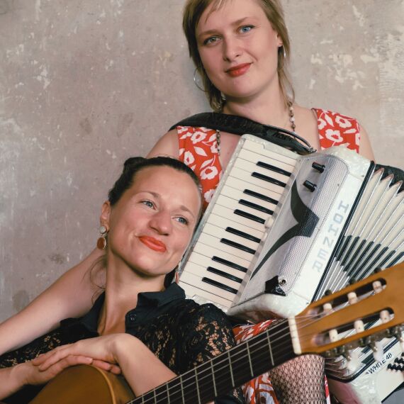 Es sind zwei Frauen mit Instrumenten zu sehen. Die Frau auf der linken Seite sitzt und hat dabei eine braune Gitarre auf dem Schoß. Ihre Hände liegen auf der Gitarre und sie schaut verträumt und mit einem Lächeln. Die Frau auf der rechten Seite steht und hat ein Akkordeon in der Hand. Sie lächelt in die Kamera.