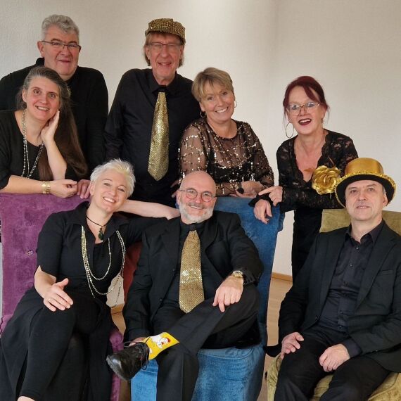 Es ist eine Band von 8 Personen zu sehen, vier Frauen und vier Männer. Alle tragen etwas Schwarzes mit goldenen Akzenten und lächeln in die Kamera. Der Mann in der Mitte trägt einen goldenen Hut und eine goldene Krawatte. Ebenso wie der Mann ganz rechts, dieser sitzt zudem auf einem goldenen Stuhl und trägt einen glänzend-goldenen Hut mit einer großer goldenen Rose integriert.  Der Mann in der Mitte sitzt auf einem blauen Stuhl und trägt goldene Socken und eine Brille. Zwei Damen auf der rechten Seite tragen eine große goldene Kette.