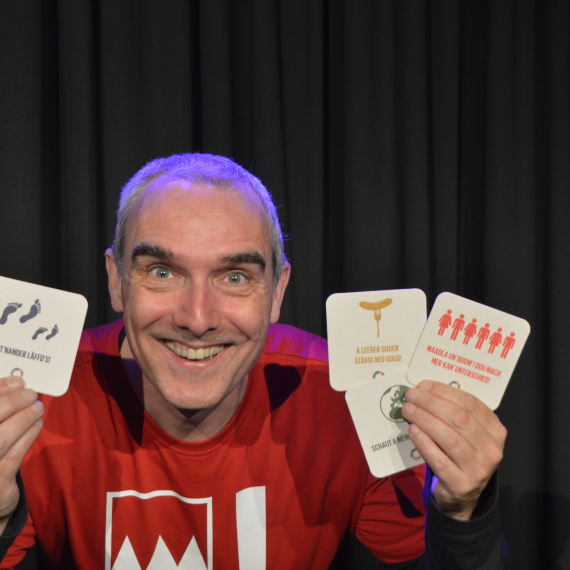 Ein Mann mit rotem T-Shirt hält vier farbige Bierdeckel in den Händen und grinst in die Kamera.