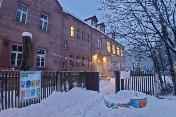 In einem großen scheebedeckten Sandsteinhaus leuchten die Fenster. Vor dem Haus hängen farbige Plakate und eine bunte Wimpelkette.