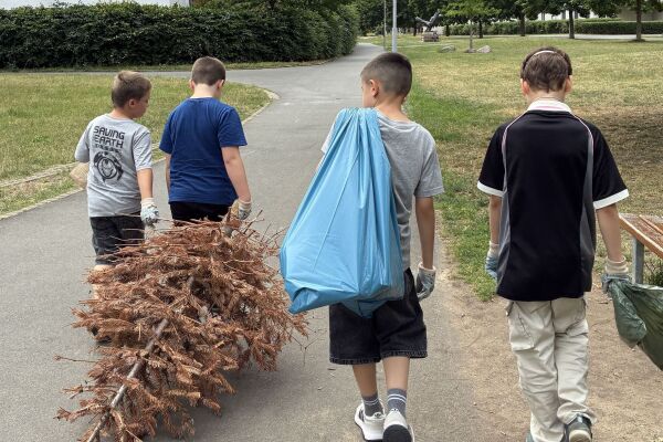 Vier Kinder laufen ausgestattet mit Müllsäcken umher, einer hat einen vertrockneten Tannenbaum im Schlepptau, während ein anderer einen blauen Müllsack trägt. Alle tragen Müllhandschuhe zum Müllsammeln.