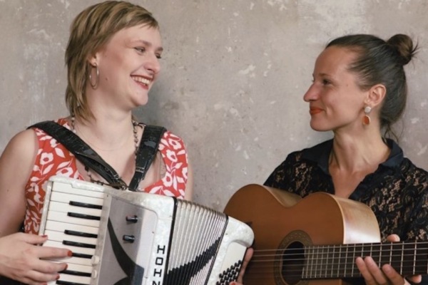 Zwei Musikerinnen, die Linke mit Akkordeon und die rechts mit Gitarre, schauen sich lächelnd an.