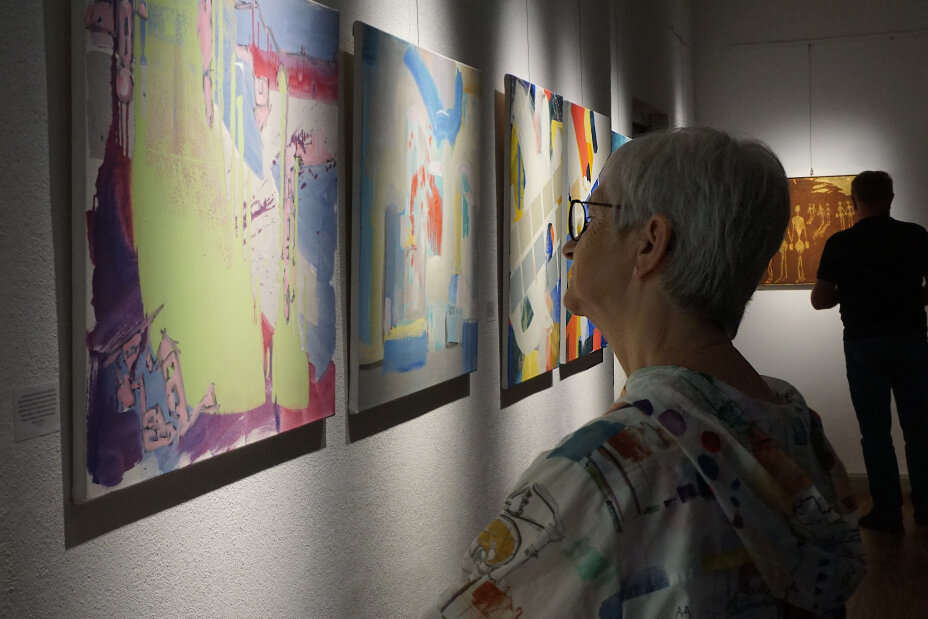 Eine Frau steht vor einer weißen Wand. Vor ihr hängt ein Gemälde des Künstlers. Sie schaut dieses vertieft an. Es handelt sich um abstrakte Kunst, bei der Pastellfarben verwendet wurden. Grün, Lila, rosa, hellblau und pink stechen heraus. Im Hintergrund hängen drei weitere Bilder. Hier wurden besonders die Farben blau, rot und gelb verwendet, Bild © Sonja Wolfstädter / Stadt Nürnberg