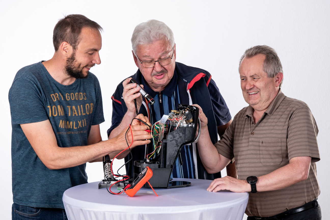 Auf einem Tisch mit weißer Tischdecke steht eine Kaffeemaschine ohne Plastikhülle. Alle Kabel sind sichtbar und drei Männer mit Schraubenzieher und Taschenlampe inspizieren das Gerät.