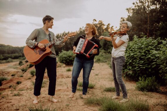 Drei Frauen stehen barfuß vor einem Wald und musizieren mit Gitarre, Akkordeon und Geige.