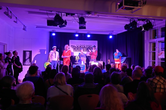 Blick von hinten auf ein sitzenden Publikum während einer Bühnenshow in einem lila beleuchteten Saal.