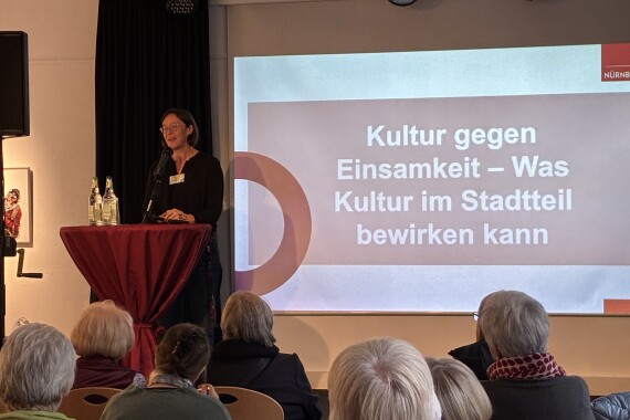 Auf einer Bühne steht eine schwarz gekleidete Frau vor einem roten Stehtisch und spricht in ein Mikrofon.