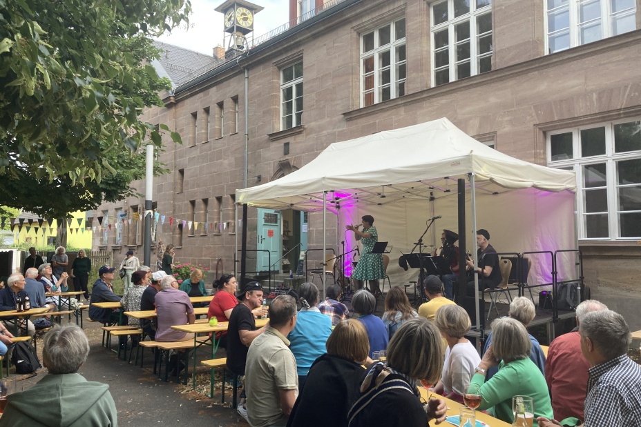 Ein Publikum sitzt an Biertischgarnituren im Freien vor einer kleinen, überdachten Bühne mit Musikern., Bild © Sonja Wolfstädter / Stadt Nürnberg