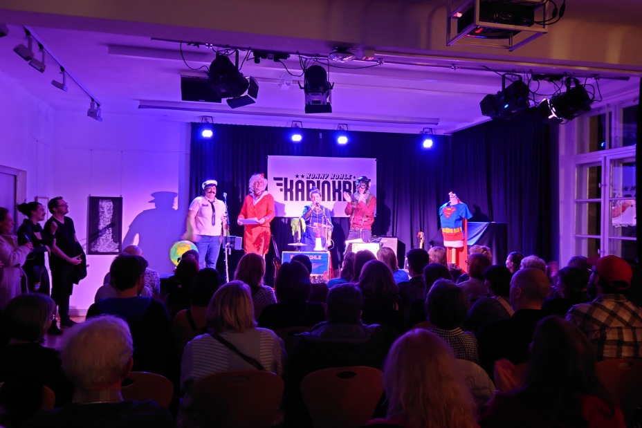 Blick von hinten auf ein sitzenden Publikum während einer Bühnenshow in einem lila beleuchteten Saal., Bild © Birgit Kretz / Stadt Nürnberg