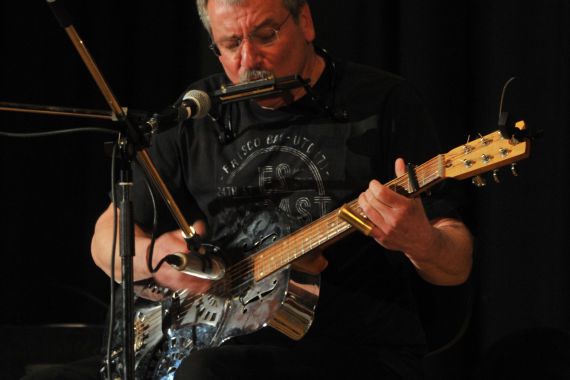 Vor schwarzem Hintergrund steht ein Mann mit kurzen grauen Haaren und schwarzem T-Shirt, er spielt Mundharmonika und Gitarre gleichzeitig.