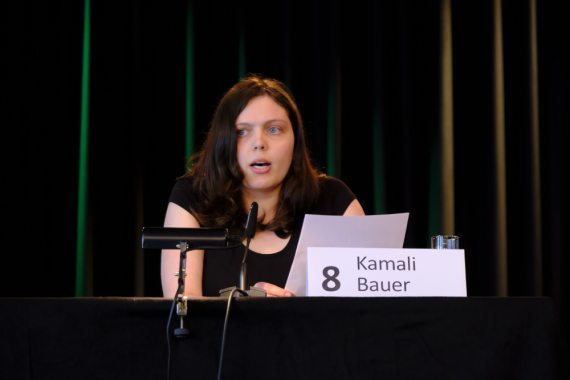 Auf einer Bühne sitzt eine junge Frau mit langen braunen Haaren und spricht in ein Mikrofon. Vor ihr steht ein Schild mit dem Namen Kamali Bauer.