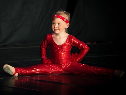Foto von einem Mädchen in einem roten Turn·anzug. Das Mädchen sitzt im Spagat auf dem Boden. Das Mädchen macht bei einem Ballett mit., Bild © Max / Sternenhaus