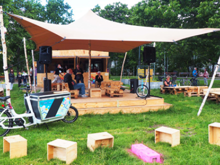 Foto vom mobilen Kultur·laden KommVorZone im Anna·park. Auf einer Wiese steht ein Zelt. Unter dem Zelt steht eine Bühne aus Holz. vor der Bühne stehen Holz·würfel zum Sitzen., Bild © Stadt Nürnberg