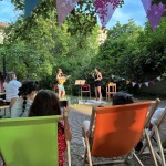 Blick über zwei Personen in Liegestühlen auf den Bühnenbereich im Grünen. Zwei Musikerinnen stehen im Hintergrund auf einer Wiese vor Bäumen und Hecken.
