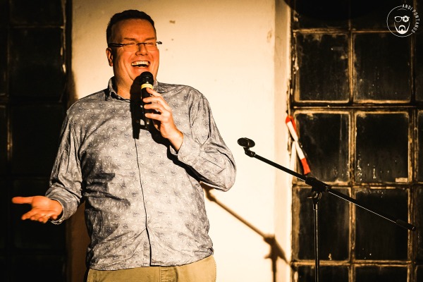 Ein Mann mit Mikrofon steht lachend vor einer weißen Wand