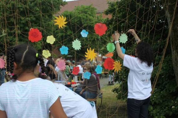 Kinder schmücken Netz mit bunten Wunschblüten. Im Hintergrund sind Büsche und Bäume.