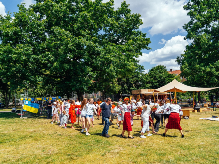 Foto von vielen Menschen auf einer Wiese. Die Menschen halten sich an den Händen und tanzen. Manche Menschen sind verkleidet., Bild © Kit Viatkin