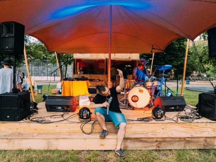 Foto von einer Bühne aus Holz. Auf der Bühne stehen viele Geräte und Instrumente verteilt. Vorne an der Bühne sitzt ein Mann mit einem Mikrofon in der Hand., Bild © Kit Viatkin