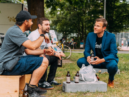 Foto von 3 Männern auf einer Wiese. 2 Männer sitzen auf niedrigen Holz·hockern. Die 2 Männer sind sportlich gekleidet. Der 3. Mann sitzt in der Hocke. Der Mann trägt einen Anzug. Die Männer halten Getränke∙flaschen in der Hand. Und die Männer unterhalten sich., Bild © Kit Viatkin