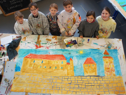 Foto von einer Gruppe von Kindern. Die Kinder haben Farb∙kleckse auf der Kleidung. Die Kinder stehen an einem Tisch. Auf dem Tisch liegt ein großes gemaltes Bild., Bild © Stadt Nürnberg