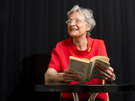 Eine Frau mit grauen Haaren und einer Brille sitzt an einem Tisch. Sie trägt ein rotes Tshirt und liest aus einem Buch, das sie in der Hand hält., Bild © Uwe Niklas / Stadt Nürnberg