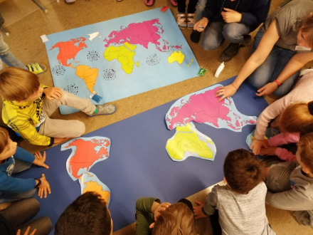 Foto von einer Gruppe von Kindern. Die Kinder sitzen um ein großes Stück blaues Papier. Auf dem blauen Papier liegen kleinere bunte Papier∙stücke. Die kleineren Papier∙stücke haben die Form von den Kontinenten., Bild © Stadt Nürnberg