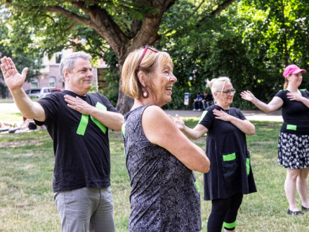 Foto von einer Gruppe von Menschen. Die Menschen stehen auf einer Wiese. Die Menschen machen alle die gleiche Bewegung: Die linke Hand liegt über der rechten Brust. Der rechte Arm ist zur Seite ausgestreckt., Bild © Olga Komarova