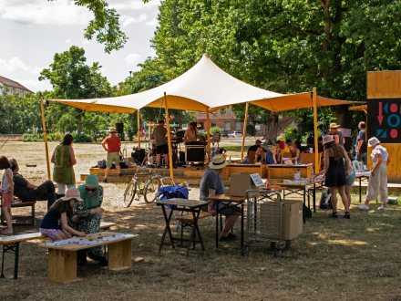 Foto von Menschen an Tischen und Bänken auf einer Wiese. Im Hintergrund ist ein Zelt·dach aufgebaut. Unter dem Zelt·dach ist eine Bühne., Bild © Harald Schmidt