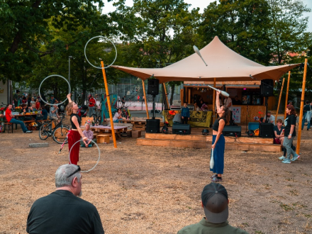 Foto von 2 Menschen auf einer Wiese. Die linke Person jongliert mit 3 Reifen. Die rechte Person jongliert mit 3 Keulen. Um die 2 Personen stehen und sitzen Menschen. Im Hintergrund ist ein Zelt∙dach aufgebaut., Bild © Kit Viatkin