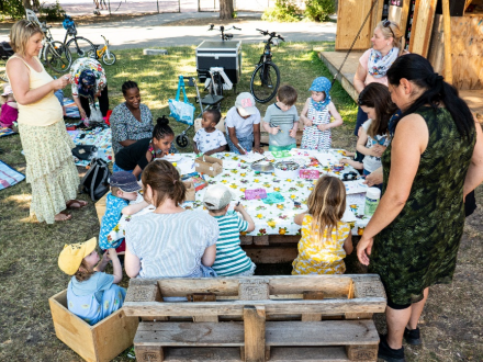 Foto von einer Gruppe von Kindern und Erwachsenen um einen Tisch herum. Die Kinder basteln. Auf der Wiese im Hintergrund liegen Picknick·decken., Bild © Harald Schmidt