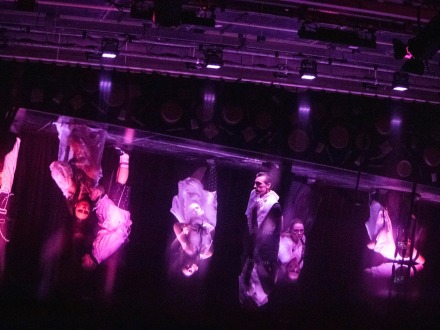 Foto von einem Theater·stück auf einer Bühne. Die Bühne ist dunkel. Die Schauspieler stehen in hellem Licht., Bild © Olga Kelsch