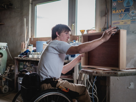 Foto von einem jungen Mann in einer Schreinerei. Der Mann schleift gerade eine Schublade aus Holz. Der Mann sitzt im Rollstuhl. © Andi Weiland / www.gesellschaftbilder.de