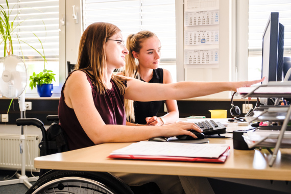 Foto von 2 Frauen an einem Schreib·tisch. Die eine Frau sitzt in einem Rollstuhl. Auf dem Schreib·tisch steht ein Computer. Die Frau im Rollstuhl zeigt der anderen Frau etwas auf dem Computer·bildschirm.