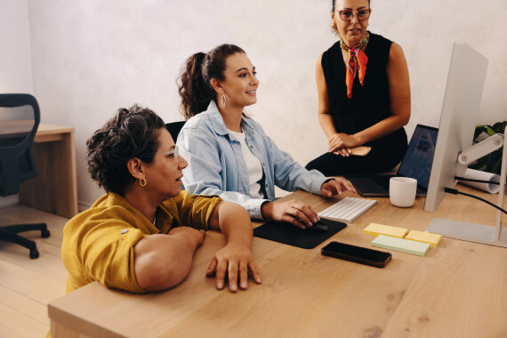 Foto von 3 Personen an einem Schreib·tisch. Die Personen besprechen etwas.